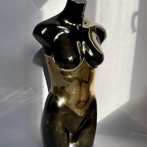 May include: A metallic sculpture of a female torso. The upper portion is black, transitioning to a gold-colored midsection and lower body. The sculpture has a smooth, reflective surface, and is set against a white background.