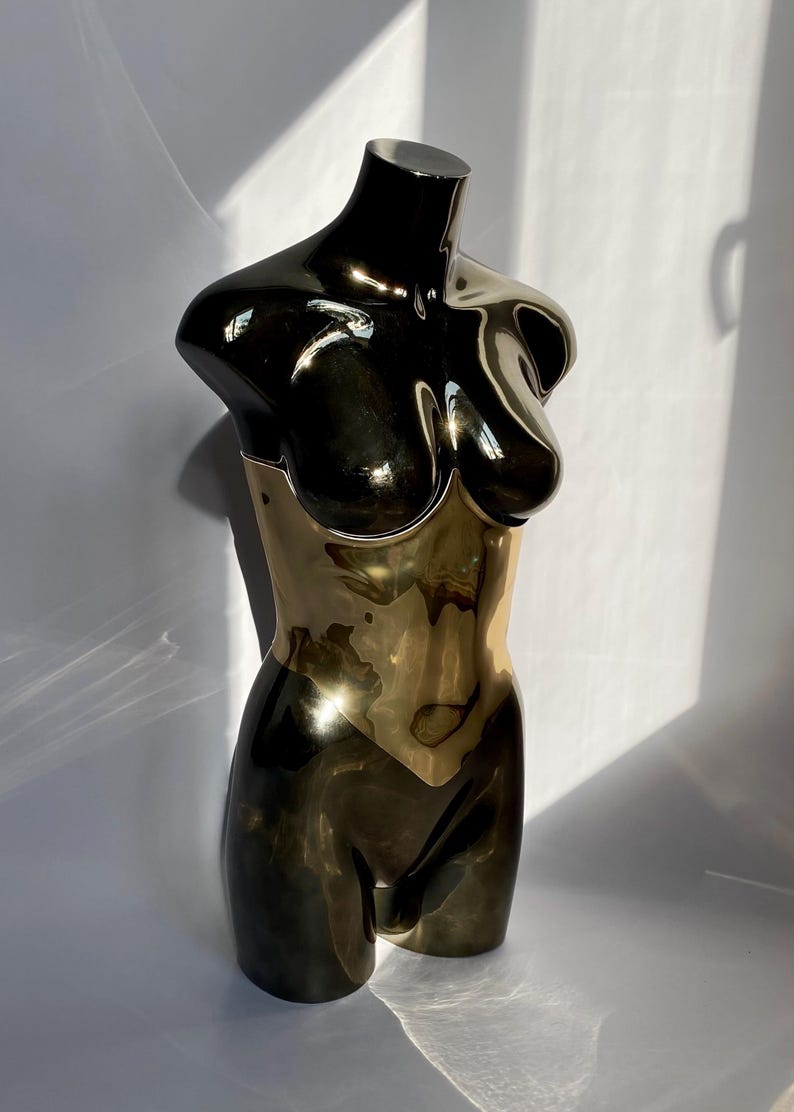 May include: A metallic sculpture of a female torso. The upper portion is black, transitioning to a gold-colored midsection and lower body. The sculpture has a smooth, reflective surface, and is set against a white background.