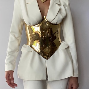 Puede incluir: Un blazer blanco combinado con un cintur&oacute;n tipo cors&eacute; de metal dorado pulido y un collar gargantilla a juego en forma de V. El blazer presenta mangas largas, solapas de muesca y dos bolsillos con solapa en la cintura, junto a pantalones blancos.