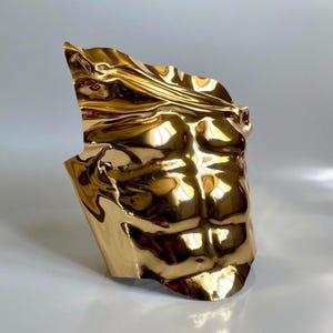 Puede incluir: Una escultura dorada de un torso, que muestra músculos abdominales definidos. La superficie metálica refleja la luz, creando reflejos y sombras. La escultura tiene una apariencia rugosa y texturizada.
