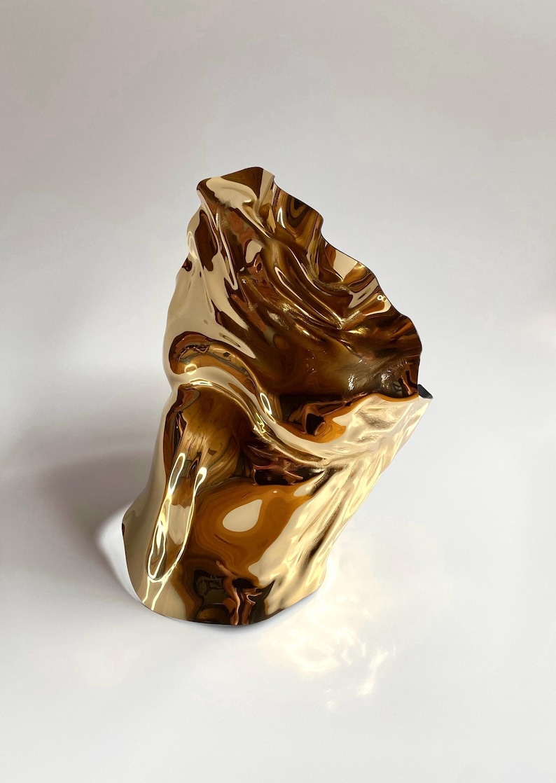 Puede incluir: Una escultura abstracta dorada con un dise&ntilde;o ondulado y fluido. La superficie met&aacute;lica refleja la luz, creando reflejos y sombras. La forma es org&aacute;nica y parece estar hecha de un material flexible.