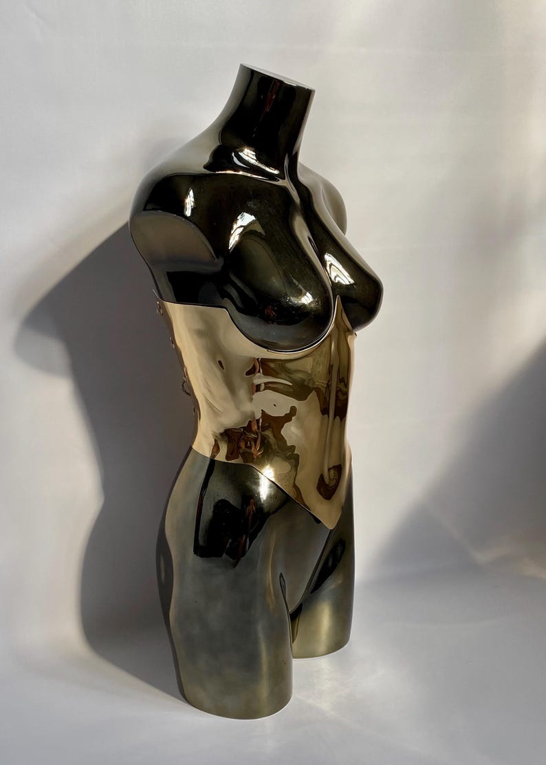 May include: A metallic sculpture of a torso, featuring a two-tone design. The upper portion is a glossy black, transitioning to a gold-colored midsection. The sculpture is a stylized representation of the human form, with smooth, reflective surfaces.