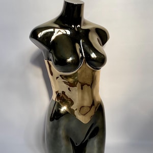 Puede incluir: Una escultura met&aacute;lica de un torso femenino. La parte superior es de un color oscuro y reflectante, que transiciona a una secci&oacute;n media dorada. La escultura tiene un acabado liso y pulido, y est&aacute; sobre un fondo blanco.