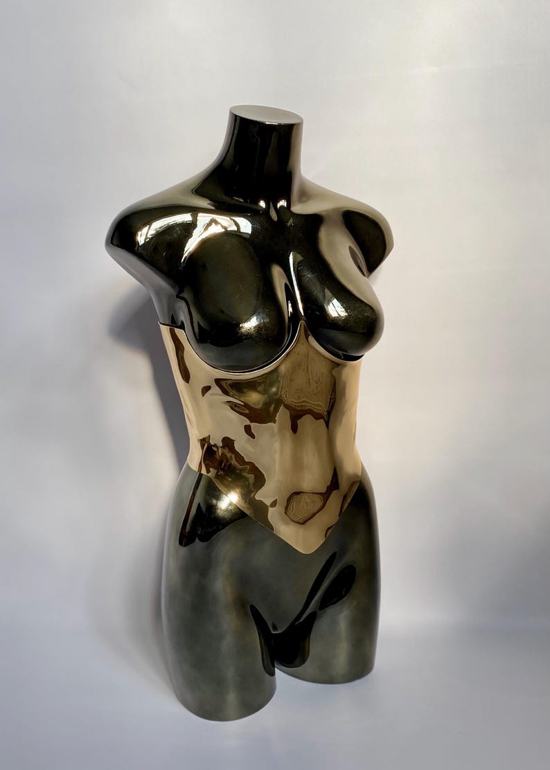 Puede incluir: Una escultura met&aacute;lica de un torso femenino. La parte superior es de un color oscuro y reflectante, que transiciona a una secci&oacute;n media dorada. La escultura tiene un acabado liso y pulido, y est&aacute; sobre un fondo blanco.