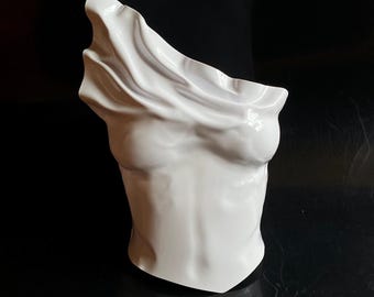 May include: A white ceramic bust sculpture depicting a torso with draped fabric over the head and shoulders. The sculpture has a smooth, glossy finish and is set against a dark background.