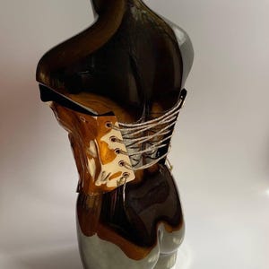 Puede incluir: Escultura de torso de cer&aacute;mica decorativa con un acabado brillante y reflectante. La parte superior es de un marr&oacute;n met&aacute;lico oscuro, que pasa a un gris plateado m&aacute;s claro en la base. Un detalle tipo cors&eacute; con cord&oacute;n plateado a&ntilde;ade una textura contrastante.