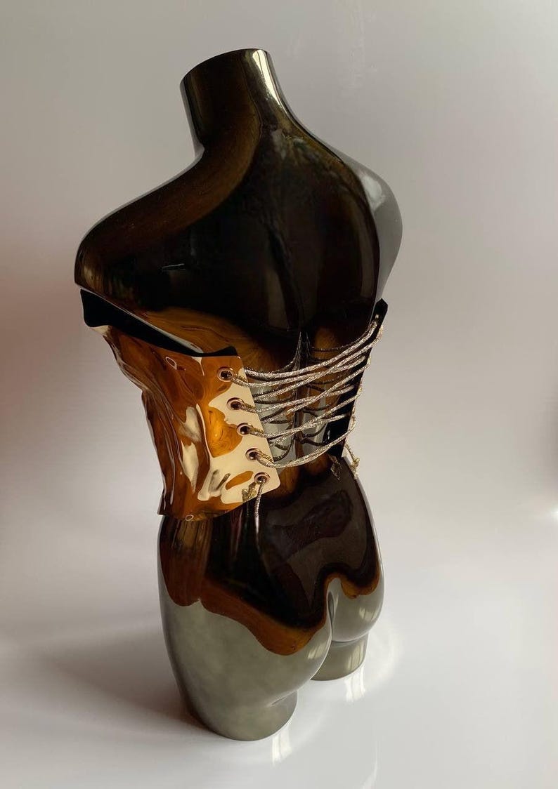 Puede incluir: Escultura de torso de cer&aacute;mica decorativa con un acabado brillante y reflectante. La parte superior es de un marr&oacute;n met&aacute;lico oscuro, que pasa a un gris plateado m&aacute;s claro en la base. Un detalle tipo cors&eacute; con cord&oacute;n plateado a&ntilde;ade una textura contrastante.