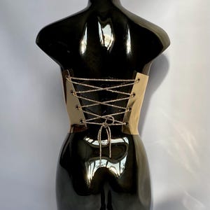 Puede incluir: Un torso de maniqu&iacute; negro con un cors&eacute; dorado. El cors&eacute; presenta un dise&ntilde;o de cordones con un cord&oacute;n plateado brillante. El maniqu&iacute; tiene un acabado brillante, que refleja la luz.