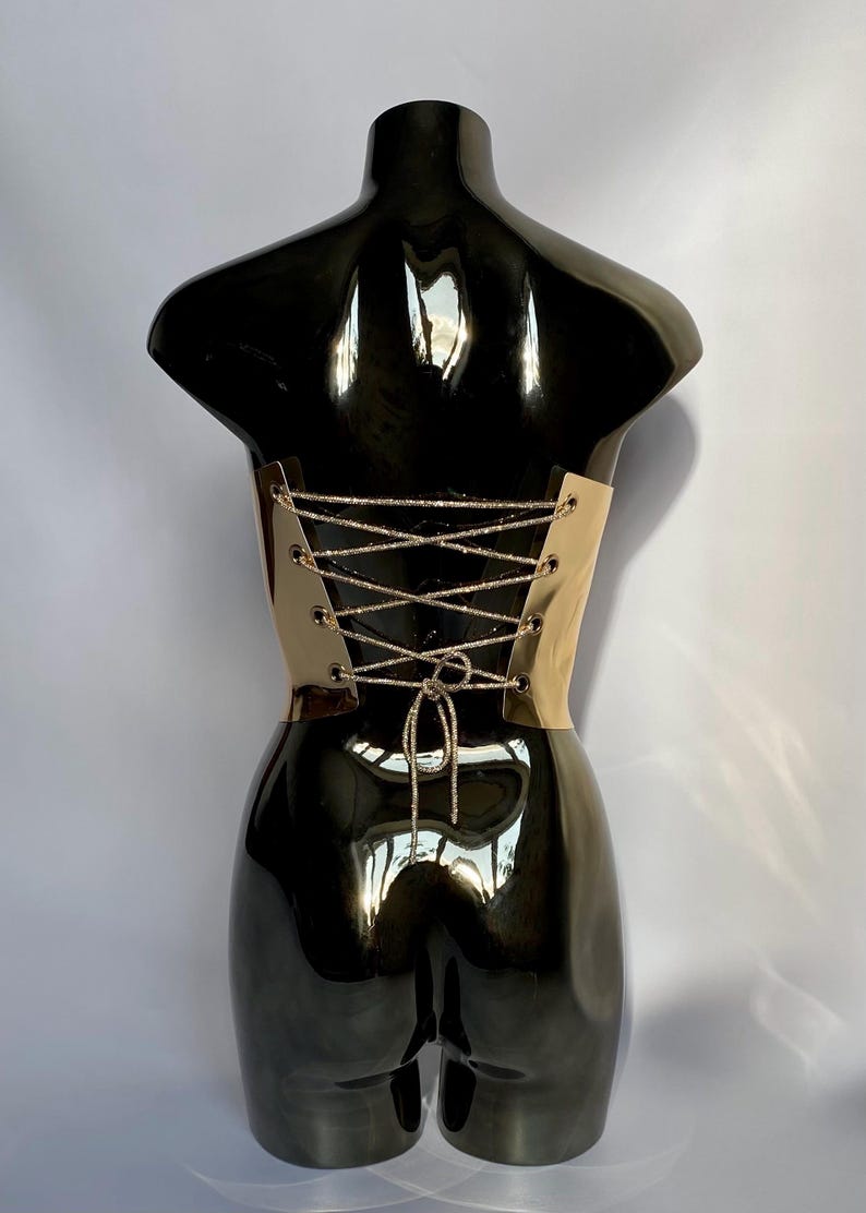 Puede incluir: Un torso de maniqu&iacute; negro con un cors&eacute; dorado. El cors&eacute; presenta un dise&ntilde;o de cordones con un cord&oacute;n plateado brillante. El maniqu&iacute; tiene un acabado brillante, que refleja la luz.