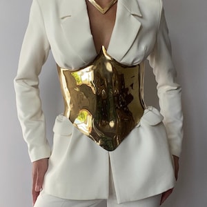 May include: Cream-colored suit featuring a gold-colored, sculpted corset. The suit includes a blazer with a notched lapel and matching trousers. A gold necklace complements the outfit. The corset appears to be made of a reflective material.