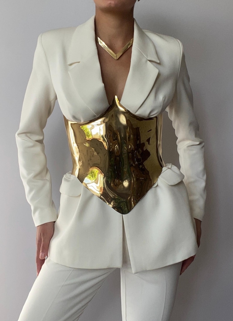 May include: Cream-colored suit featuring a gold-colored, sculpted corset. The suit includes a blazer with a notched lapel and matching trousers. A gold necklace complements the outfit. The corset appears to be made of a reflective material.