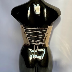May include: A beige corset with a lace-up back is displayed on a black mannequin. The corset has a metallic sheen and is laced with silver-colored cord. The mannequin is glossy and reflects the surrounding environment.