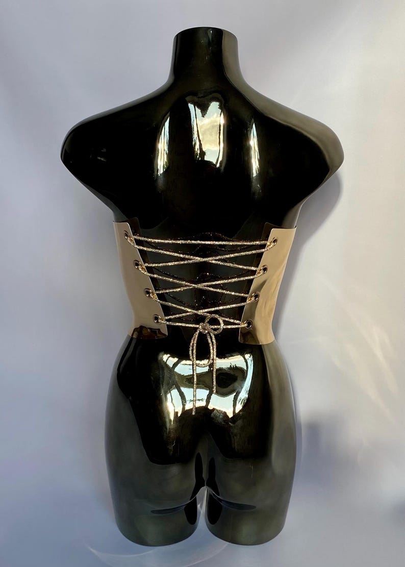 May include: A beige corset with a lace-up back is displayed on a black mannequin. The corset has a metallic sheen and is laced with silver-colored cord. The mannequin is glossy and reflects the surrounding environment.
