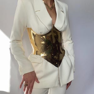 Puede incluir: Un blazer color crema y pantalones a juego combinados con un cors&eacute; de metal dorado. El blazer tiene un dise&ntilde;o cl&aacute;sico con solapas y bolsillos. El cors&eacute; es una pieza llamativa, que a&ntilde;ade un contraste audaz al conjunto. Un collar dorado completa el look.