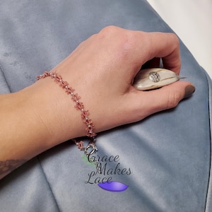 May include: A delicate pink crocheted bracelet with a silver clasp. The bracelet is made with small, intricate stitches and has a delicate, feminine look. The bracelet is shown on a person's wrist.