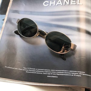 May include: Oval-shaped sunglasses with a metallic silver frame and dark, reflective lenses. The sunglasses are positioned on a page featuring the text J 12 CHANEL and additional Russian text describing a watch, including details about titanium ceramic.