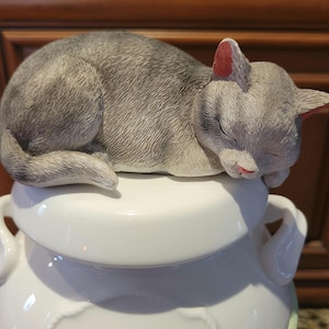 Pode incluir: Um pote de biscoitos de cerâmica branca com uma figura de gato dormindo cinza e branco na tampa. O gato tem orelhas cor-de-rosa e um nariz cor-de-rosa.