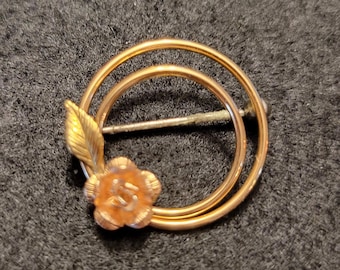 Peut inclure: Une broche dorée représentant une rose stylisée avec une feuille, placée dans un double cercle.