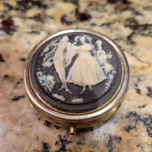 Puede incluir: Un pastillero redondo y antiguo con un marco de metal dorado. La tapa presenta un camafeo en blanco y negro de una pareja bailando. El camafeo está rodeado por un borde de metal dorado.