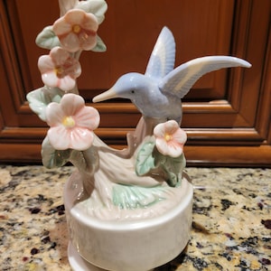 Puede incluir: Figura de colibrí de cerámica en azul claro y blanco, posada en una rama con flores rosas y hojas verdes. La figura se asienta sobre una base redonda blanca, probablemente una caja de música. El fondo es un mueble de madera.