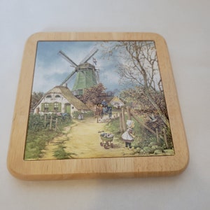Peut inclure: Un dessous de plat en bois avec une tuile en céramique représentant une illustration colorée d'un moulin à vent hollandais, un petit village et une fille avec une poussette de poupée. La tuile porte le texte "1851" sur un bâtiment.