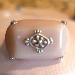 May include: A rectangular, light pink ring with a silver floral design in the center. The ring has silver accents on each corner. The ring is a piece of jewelry.