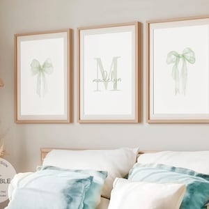 Może przedstawiać: Trzy oprawione wydruki z zieloną akwarelową kokardką, zieloną akwarelową literą M z imieniem "Madelyn" i kolejną zieloną akwarelową kokardką. Ramy są z jasnego drewna, a tło to jasnobeżowa ściana.