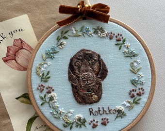 May include: Embroidered dog portrait in a wooden hoop. The dog is brown with a small gold tag. The background is light blue, surrounded by embroidered flowers and paw prints. A brown velvet bow is tied at the top.