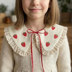 May include: A cream-colored collared shirt with a strawberry pattern. The collar is adorned with embroidered red strawberries and a delicate lace trim. A red ribbon is tied in a bow at the neckline. The shirt is part of a sweater.