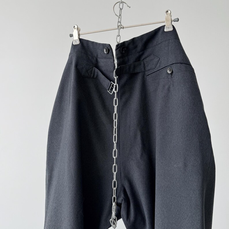 May include: Dark gray high-waisted pants hanging from a metal hanger. The pants have a button closure, belt loops, and a silver chain detail running down the front. The fabric appears to be a woven material.