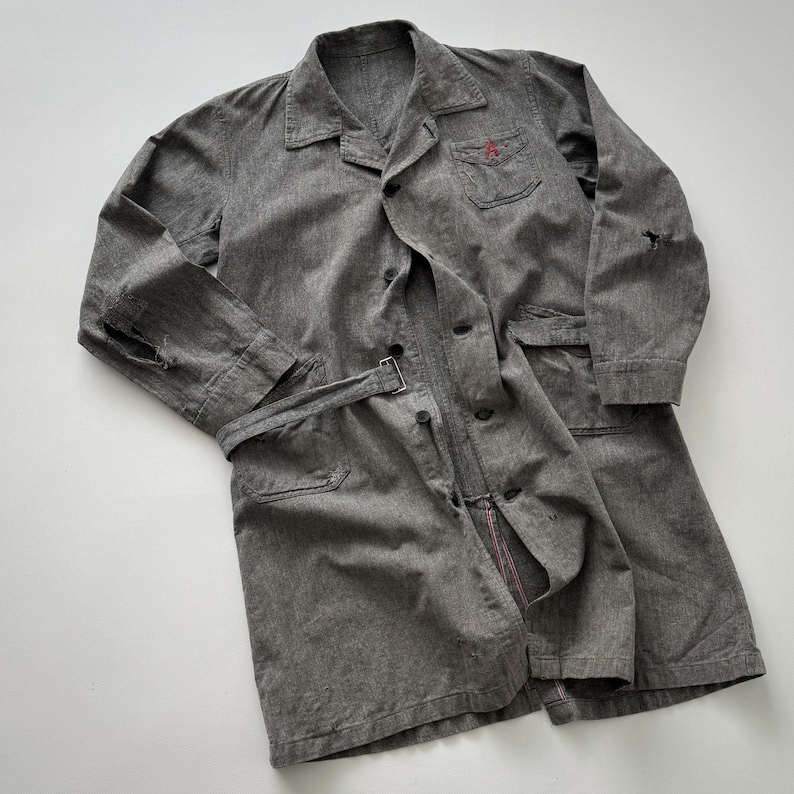 May include: A gray, long-sleeved coat with a button-up front, belt, and pockets. The coat has a small red embroidered detail on the chest pocket and a distressed look with a tear on the sleeve. The coat is made of a textured fabric.