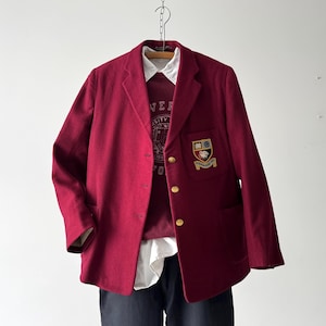 Puede incluir: Una chaqueta de color granate con botones dorados y un parche con escudo en el pecho izquierdo. La chaqueta tiene dos bolsillos en la parte delantera y un cierre de botón único.