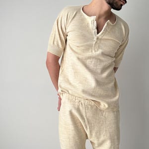 Könnte beinhalten: Ein hellbeige gestricktes Henley-Shirt mit kurzen Ärmeln und passender Hose. Das Shirt hat einen Knopfleisten-Ausschnitt. Das Outfit ist aus einem weichen, strukturierten Stoff gefertigt. Der Gesamtstil ist lässig und bequem.