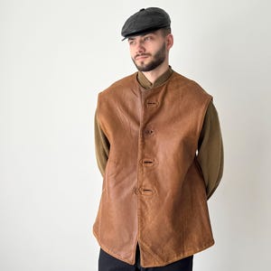 May include: A brown leather waistcoat with a button fastening. The waistcoat has a stand-up collar and a relaxed fit.