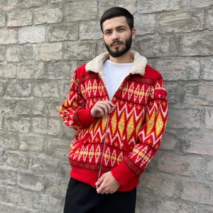 Puede incluir: Una chaqueta roja con cremallera y cuello afelpado color crema. El suéter presenta un patrón geométrico en amarillo, blanco y negro. La chaqueta es de punto.