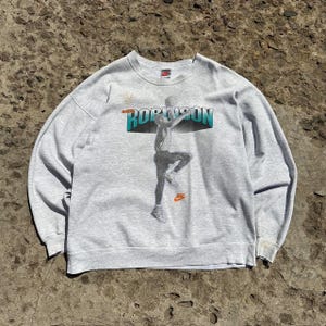 Puede incluir: Sudadera gris con un estampado gr&aacute;fico de un jugador de baloncesto en acci&oacute;n. El texto "Dennis Rodman" est&aacute; impreso en la parte delantera de la sudadera.