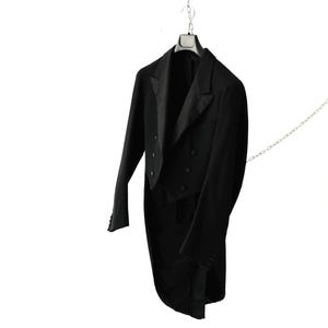 Op de afbeelding: Een zwarte rokkostuumjas met een dubbele rij knopen, satijnen revers en knopen, hangend aan een metalen hanger en een zilverkleurige ketting. Het kledingstuk heeft lange mouwen met knoopmanchetten en een gestructureerde, formele snit.