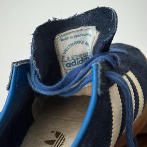 May include: Close-up of a vintage navy blue Adidas sneaker. The shoe features white stripes, blue laces, and a label that reads "Made in Yugoslavia." The Adidas logo is visible on the insole. The shoe's design is classic and retro.