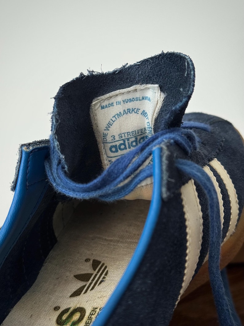 May include: Close-up of a vintage navy blue Adidas sneaker. The shoe features white stripes, blue laces, and a label that reads "Made in Yugoslavia." The Adidas logo is visible on the insole. The shoe's design is classic and retro.