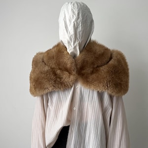 May include: A brown fur collar is draped over a striped shirt and a white hood. The collar is a warm, light brown color and appears to be made of soft fur. The shirt has vertical stripes in a light pink color.
