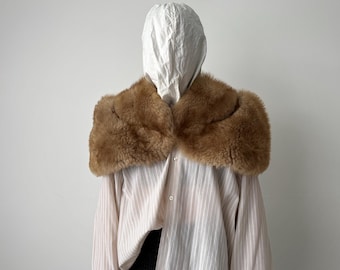Può includere: Un colletto di pelliccia marrone è drappeggiato su una camicia a righe e un cappuccio bianco. Il colletto è di un caldo marrone chiaro e sembra essere fatto di morbida pelliccia. La camicia ha righe verticali rosa chiaro.