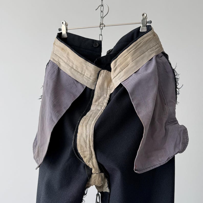 May include: Black denim pants with a unique design, featuring a light beige waistband and accents, and gray fabric panels. The pants are suspended from a metal hanger, showcasing their unusual construction and style.