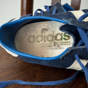 May include: Close-up of a vintage navy blue Adidas sneaker. The shoe features white stripes and blue laces. The insole displays the Adidas logo and text. The shoe is worn and shows signs of age.