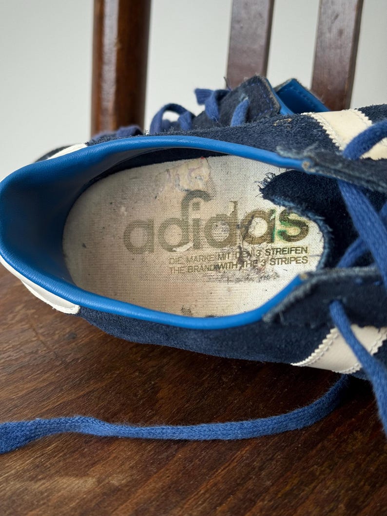 May include: Close-up of a vintage navy blue Adidas sneaker. The shoe features white stripes and blue laces. The insole displays the Adidas logo and text. The shoe is worn and shows signs of age.