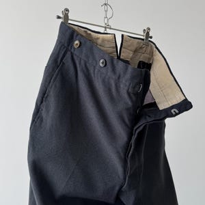 May include: Dark gray trousers hanging from a metal hanger. The pants have a button closure and a textured interior lining. The fabric appears to be a woven material. The trousers are displayed against a plain white background.