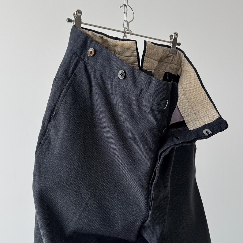 May include: Dark gray trousers hanging from a metal hanger. The pants have a button closure and a textured interior lining. The fabric appears to be a woven material. The trousers are displayed against a plain white background.