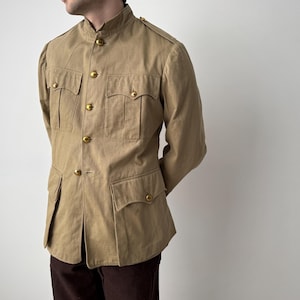 May include: Tan military-style jacket with a mandarin collar and multiple pockets. The jacket features gold-colored buttons and epaulettes on the shoulders. The long-sleeved jacket is paired with dark brown pants.