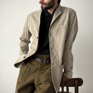 May include: A light tan blazer with a black shirt and olive green pants. The blazer has a classic design with a notched lapel and button closure. The pants have large pockets. A brown leather belt is visible.