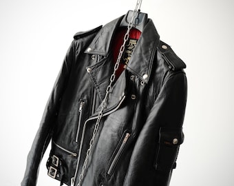 Könnte beinhalten: Eine schwarze Motorradjacke aus Leder mit leuchtend rotem Innenfutter, silberfarbenen Metallreißverschlüssen, Revers mit Druckknöpfen, Schulterklappen und verstellbaren Seitenschnallen. Eine silberne Metallkette hängt vertikal vor der Jacke.