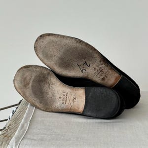 Pu&ograve; includere: Primo piano delle suole di un paio di mocassini in pelle scamosciata nera. Le suole sono in pelle marrone con il testo "GUCCI made in Italy" e la taglia "7 1/2" scritta su di esse.